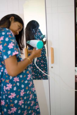 Bir kadın evinde aynanın önünde dikilirken elbiselerini ütülüyor. O, işine odaklanmış durumda..