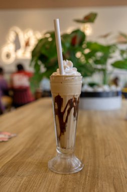 A tall glass filled with a milkshake and topped with cream sits on a table. People are in the background.