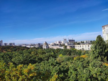 Sonbaharda modern binalar ve yeşil parklarla panoramik şehir silueti. Parlak mavi gökyüzü, renkli ağaçların üzerinde bulutlar ve gün ışığında kentsel manzara..