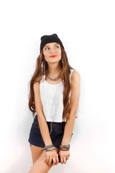 Hermosa mujer con sombrero negro y camiseta blanca escuchando música cerca de la pared blanca, sosteniendo un teléfono celular en la mano — Foto de Stock