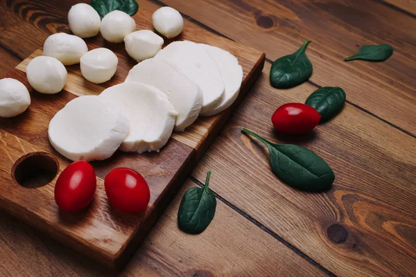Białe małe kulki serowe mozzarelli, liście szpinaku i pomidory na drewnianej desce. — Zdjęcie stockowe