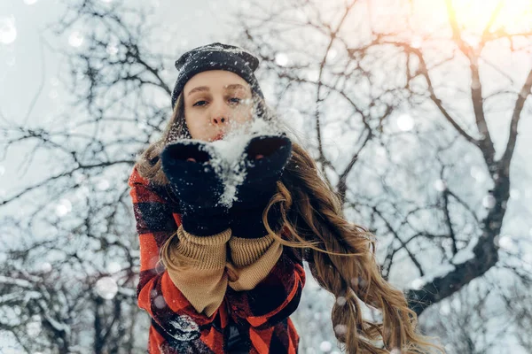 Woman Wearing Warm Winter Clothes And Hat Blowing Snow In Winter Park. Flying Snowflakes. Sunny day. Joyful Beauty young girl Having Fun in frosty Forest. — Stock Photo, Image