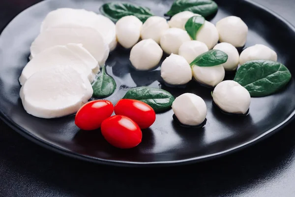 Beyaz küçük mozarella peynir topları, ıspanak yaprakları ve siyah tabakta domatesler.. Telifsiz Stok Imajlar