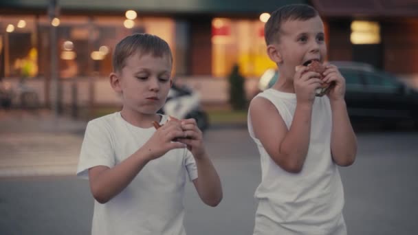เด็กชายสองคนหิวโหย กินเบอร์เกอร์บนถนนตอนเย็น . — วีดีโอสต็อก