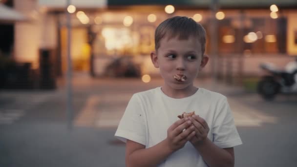 Hungriger kleiner Junge isst einen Burger auf der Abendstraße. — Stockvideo