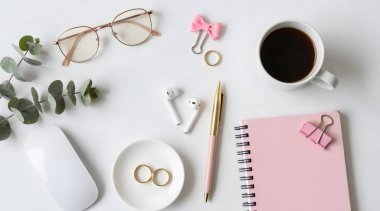 Not defteri, bardaklar, kahve fincanı, çiçekler ve pembe arka plandaki dekorasyonlarla çalışma alanının üst görüntüsü
