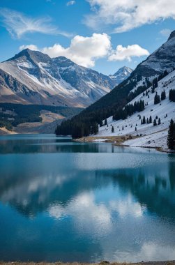 Güzel mavi bir göl karla kaplı dağlarla çevrili parlak mavi bir gökyüzünün altında tüylü beyaz bulutlarla