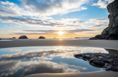 Sahilde güzel bir gün batımı. Su üzerinde kayalar yansıyor.
