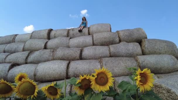Jong meisje zit op de top van de ronde hooibaal in het zonnebloemveld in de zomer in Wit-Rusland. Generatie Z. Langzame beweging. — Stockvideo