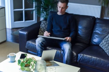 Oturma odasındaki masadan tablet ve not defteriyle çalışan bir adam.