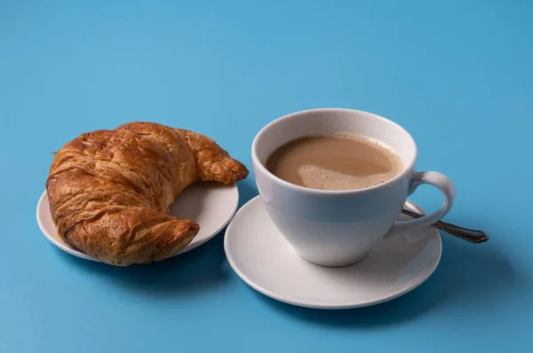 Cup of milk coffee and croissant on blue background, copy space for text.
