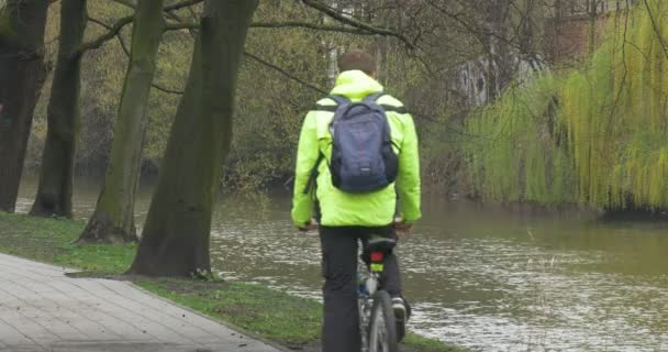 Man rijdt een fiets stopt Gets Off in Park Alley Backpacker in glazen is op zoek bij Water in de rivier hebben een Rest Water stroomt voorjaar bewolkte dag — Stockvideo