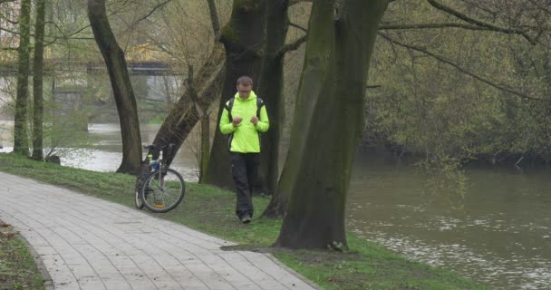 Człowiek Scrolls Tablet w pobliżu rower w parku turystycznych jest jazda rower siedzi na rowerze w parku aleja Backpacker w pobliżu rzeki wody przepływa wiosna zachmurzony — Wideo stockowe