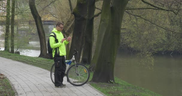 Man is een fiets in Park Alley Backpacker in de buurt van de rivier Water stroomt voorjaar bewolkt — Stockvideo