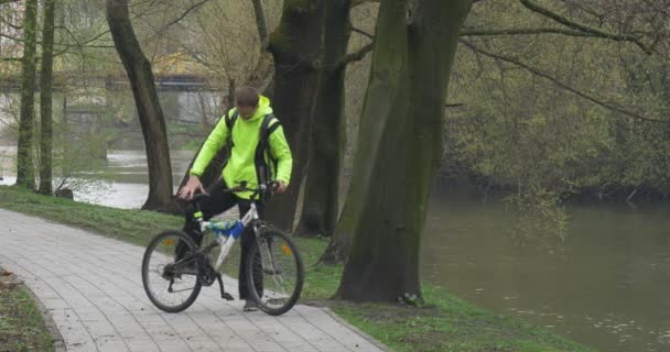 Homem está montando uma bicicleta no parque Alley Backpacker Perto do rio Fluxos de água Primavera Nublado — Vídeo de Stock