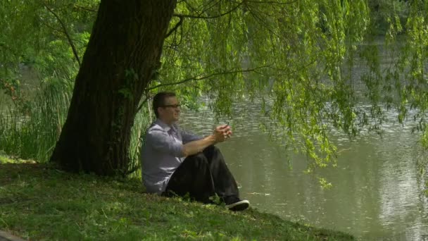 Ο άνθρωπος που κάθεται κοντά στο νερό με το κινητό τηλέφωνο κάτω από το δέντρο λίμνη Τράπεζα άνθρωπος που κάθεται σε ένα έδαφος κρατώντας ένα Smartphone φρέσκα πράσινα δέντρα ηλιόλουστη καλοκαιρινή μέρα — Αρχείο Βίντεο