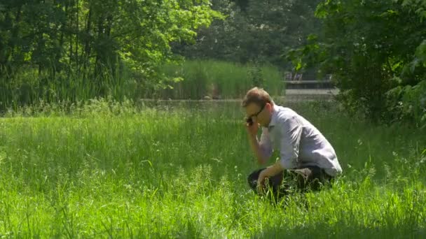 Человек, говорящий по телефону сидит на земле зеленый парк делает видео волн рукой говорит привет Щелкает смартфон провести время на природе рядом с озером — стоковое видео