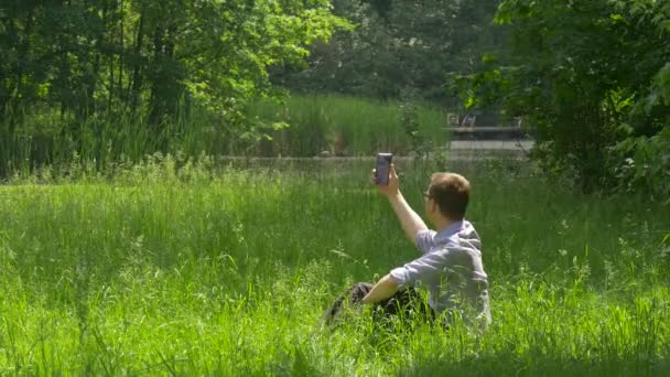 Un homme tenant un téléphone portable prend des photos dans un parc Faire un vidéocall Un homme se lève pour une promenade au sol Passez du temps à la nature Arbres verts frais Ensoleillé Été — Video