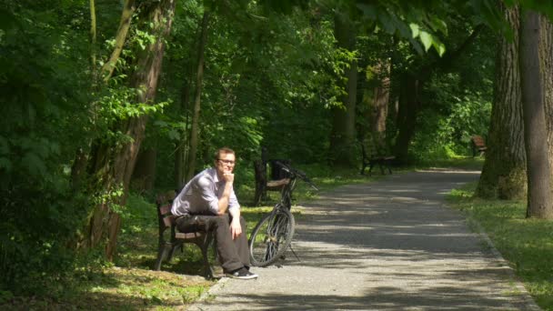 公園を考えて自転車でベンチに座っている男が左公園の路地の近く、ベンチの下で木観光では彼の腕がある残り夏晴れた日に身を乗り出して — ストック動画