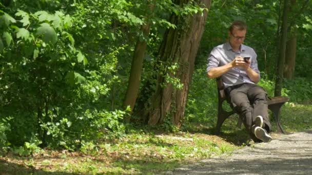 Mann sitzt mit Handy im grünen Park und plaudert auf einer Bank sitzend mit gestreckten Beinen Zeit in der Natur verbringen frische grüne Bäume sonniger Sommertag — Stockvideo