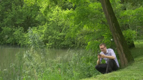 Man Clicks Mobile Phone Under Acacia Gets up Lake River Bank Man is Sitting Near the Water Holding a Smartphone Fresh Green Trees Sunny Summer Day — Stock Video