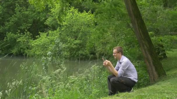 Άνθρωπος κλικ με εικονικό δισκίο στο πάρκο Διαδίκτυο αγορές εργάζονται για Forex μηνύματα άνθρωπος είναι καθισμένος κοντά στο νερό φρέσκα πράσινα δέντρα ηλιόλουστη θερινή ημέρα — Αρχείο Βίντεο