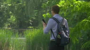 Bir cep telefonu turist adam doğa taze yeşil ağaçlar güneşli gün backpacker ile Smartphone O'nun geri kamera yaz Park Holding için yürüyor