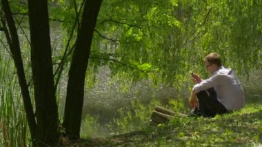 Cep telefonu oturur bir ileti numarası yazarak Park zemin Lake bankaya olan adam harcamak zaman taze yeşil ağaçlar Willow güneşli yaz günü altında Nature