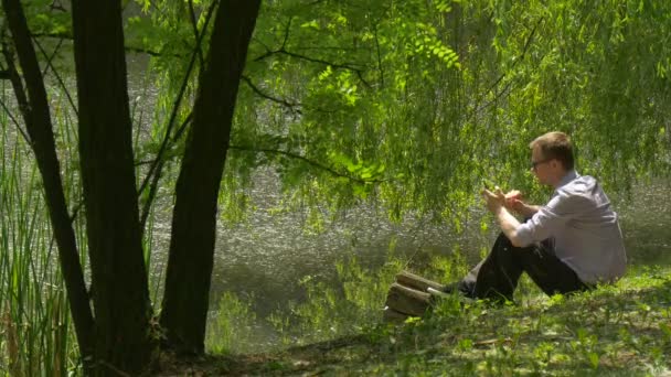 Mann tippt eine Nummer auf Handy, sitzt am Ufer des Sees im Park und redet Zeit in der Natur verbringen frische grüne Bäume Weiden sonnigen Sommertag — Stockvideo