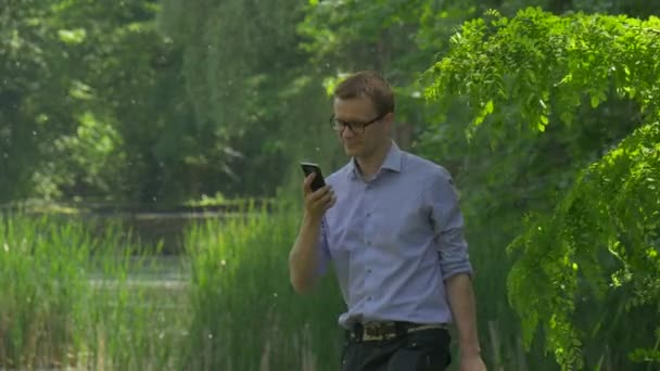 Man Types a Number Talking on Smartphone in Park Clicks Telefone móvel Passe um tempo na natureza Árvores verdes frescas Lago Acacia álamo Algodão ensolarado Dia de Verão — Vídeo de Stock