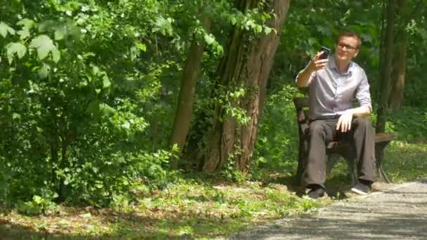 Hombre haciendo videollamada ondea su teléfono móvil de mano en Green Park sosteniendo un teléfono inteligente mirando la cámara Pasa tiempo entre árboles verdes Soleado día de verano — Vídeos de Stock