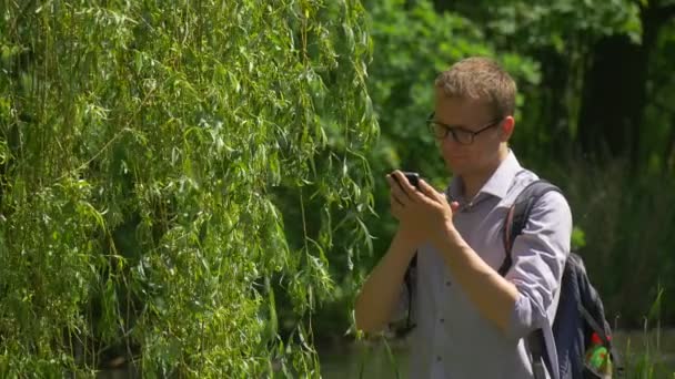 Muž v brýlích klepne telefon v parku odcházet, že batohem tráví čas v přírodě svěží zelené stromy jezero vody Willow větve slunečný letní den — Stock video