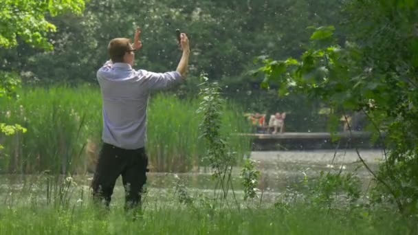 Man Waves his Hand into Mobile Phone in Park Man Making Videocall Near the Lake Holding a Smartphone Taking Selfie Spend Time at the Nature Summer Day — стоковое видео