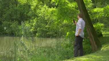 Dostum gelir Gölü banka gülümseyen adam gözlüklü telefonla konuşmayı suyun kenarında bir Smartphone taze yeşil ağaçlar güneşli yaz günü Holding duruyor