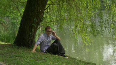 Dostum Tıklanma cep telefonu arama banka Lake alır uzak yürüyor dostum bir Smartphone taze yeşil ağaçlar güneşli yaz günü Holding su kenarında oturuyor