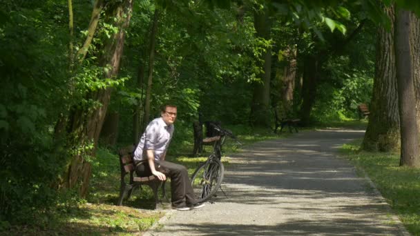 Man Takes Backpack Off Sits on Bench Park Bicicleta é deixada em Park Alley Perto do banco em Park Alley Banco Sob Árvores Turista Descanse Verão Dia ensolarado — Vídeo de Stock