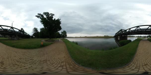 360vr wideo człowieka idącego przez nawierzchni kamieni River Bank zielona trawa samochodów na moście przez rzekę młody człowiek z plecak turystyczny w pochmurny dzień falujący wody — Wideo stockowe