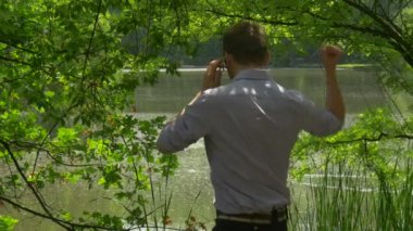 Yumruğunu diyor telefon yükseltir üzerinde konuşan adam göl banka Green Park üzerinde ayakta tutan bir Smartphone doğa taze yeşil ağaçlar Reed güneşli yaz günü Evet