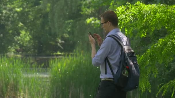 Backpacker clique sur un écran de Smartphone dans le parc tenant un téléphone portable tourne autour de l'homme marche à la nature fraîche arbres verts ensoleillé journée d'été — Video