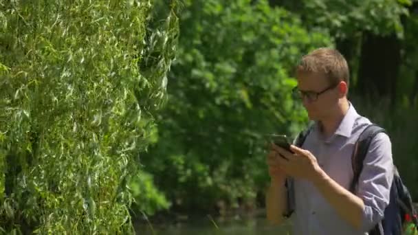 Man Clicks Telefone móvel no parque Olhando ao redor Backpacker passa o tempo na natureza Árvores verdes frescas Lago água salgueiro Ramos dia ensolarado de verão — Vídeo de Stock