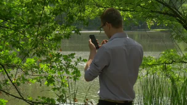 Hombre hace clic en el teléfono móvil sonriendo de pie en Lake Bank Green Park celebración de un Smartphone pasar tiempo en la naturaleza árboles verdes frescos caña soleado día de verano — Vídeos de Stock