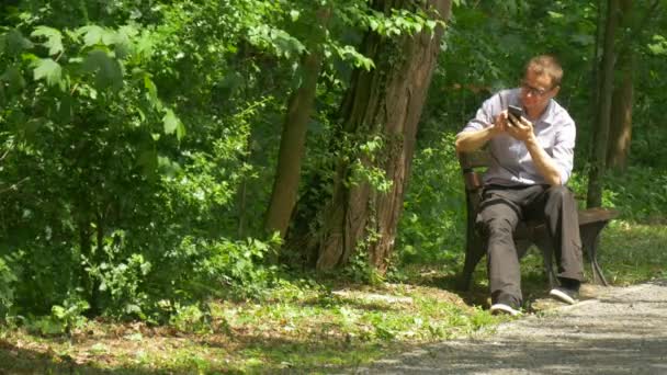 Homem com telefone móvel no parque verde bate-papo mensagens gira enquanto sentado em um banco passar o tempo na natureza no beco Árvores verdes frescas Dia ensolarado de verão — Vídeo de Stock