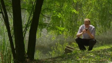 Adam göl Bankası Park kulağını yakınındaki bir akıllı telefon tutarak çömelme arama cep telefonunuzla harcamak zaman doğa taze yeşil ağaçlar güneşli yaz gün