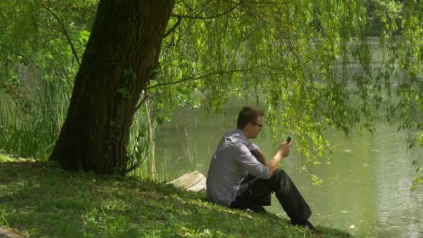 Man is Calling on Mobile Phone Under the Tree Lake Bank Man está sentado perto da água segurando um smartphone árvores verdes frescas ensolarado verão dia ondulação — Vídeo de Stock