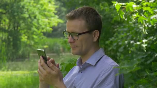 Hombre hace clic en el teléfono móvil en Park Smiling Texting lo pone cerca de su oído sentado entre árboles verdes Pasa tiempo en la naturaleza Árboles verdes frescos Verano soleado — Vídeo de stock