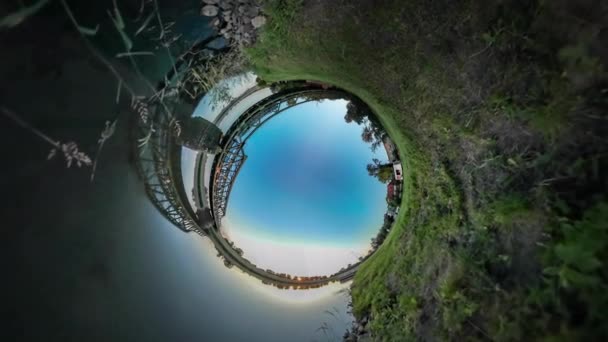 Il treno video 360Vr sta cavalcando il fiume dal villaggio delle case del ponte su un ponte di metallo della banca Il cemento sostiene i pendii verdi Pietre su un cielo blu poco profondo Estate — Video Stock