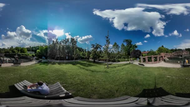 Vidéo 360Vr L'homme sur une chaise longue dans le parc Les gens marchent avec les enfants dans le jardin botanique Zoo Amusez-vous en plein air Parc allée Arbres verts ensoleillé ciel bleu 
