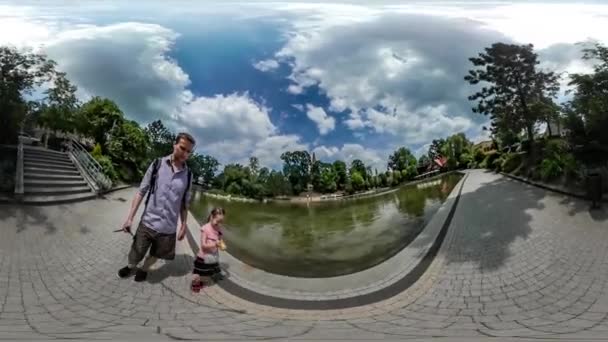 360Vr Vídeo Papá y niño en el callejón Estanque en el parque de la ciudad El hombre está sosteniendo una cámara en un palillo Familia caminando por adoquines La gente juega árboles verdes Verano — Vídeos de Stock