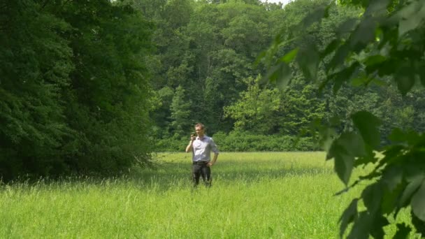 Mann mit Smartphone spaziert durch hohen grasgrünen Park Mann hält ein Telefon eine Nummer eintippt und ruft grüne üppige Bäume wachsen sonnigen Sommertag Wald — Stockvideo