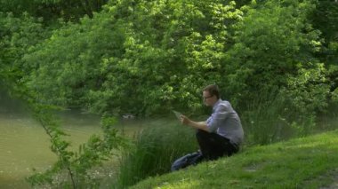 İşadamı cep telefonunuzla yeşil tepede oturan bir akıllı telefon tutarak zamanında Park taze yeşil çim ağaçlar güneşli yaz günü geçirdiği